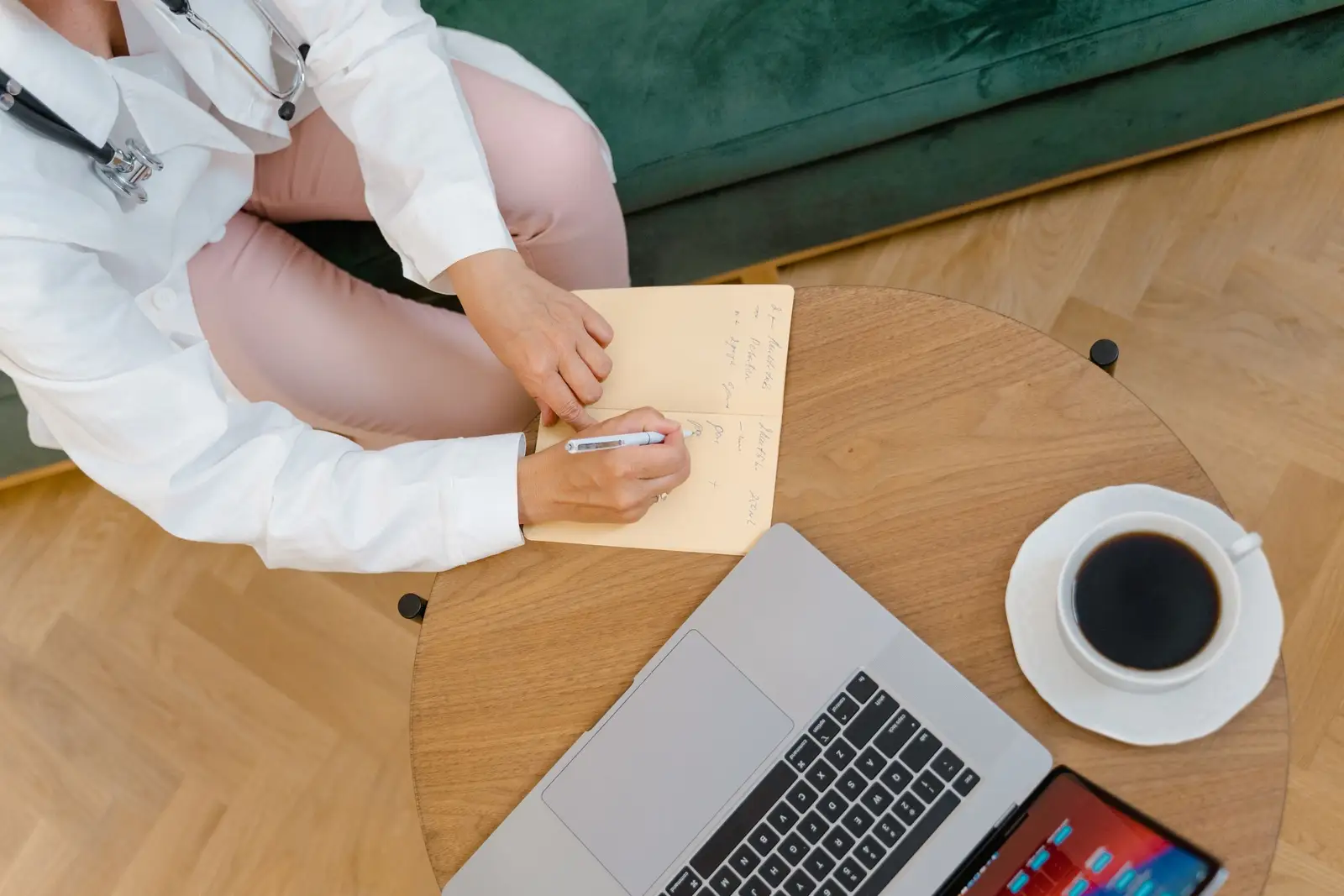 A doctor's hands writing a note in a journal on a wooden table, beside a laptop and a cup of coffee