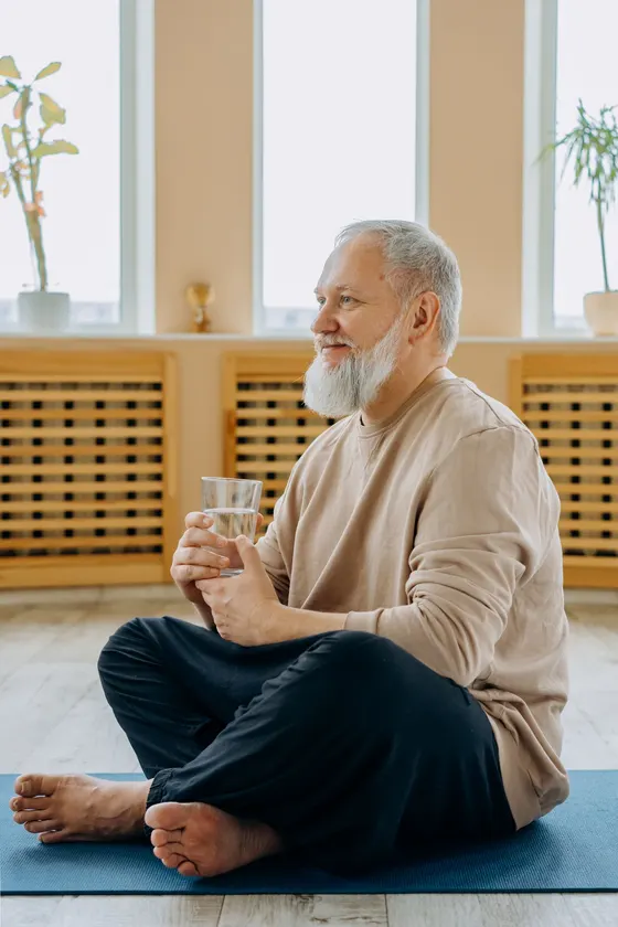 An older man sitting cross-legged in warm morning light, holding a glass of water