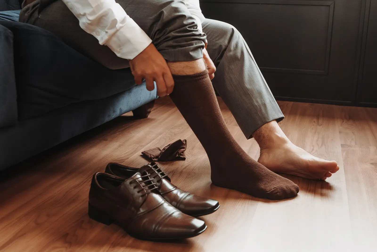 A person pulling on a knee-high sock while sitting beside a pair of shoes on a wooden floor