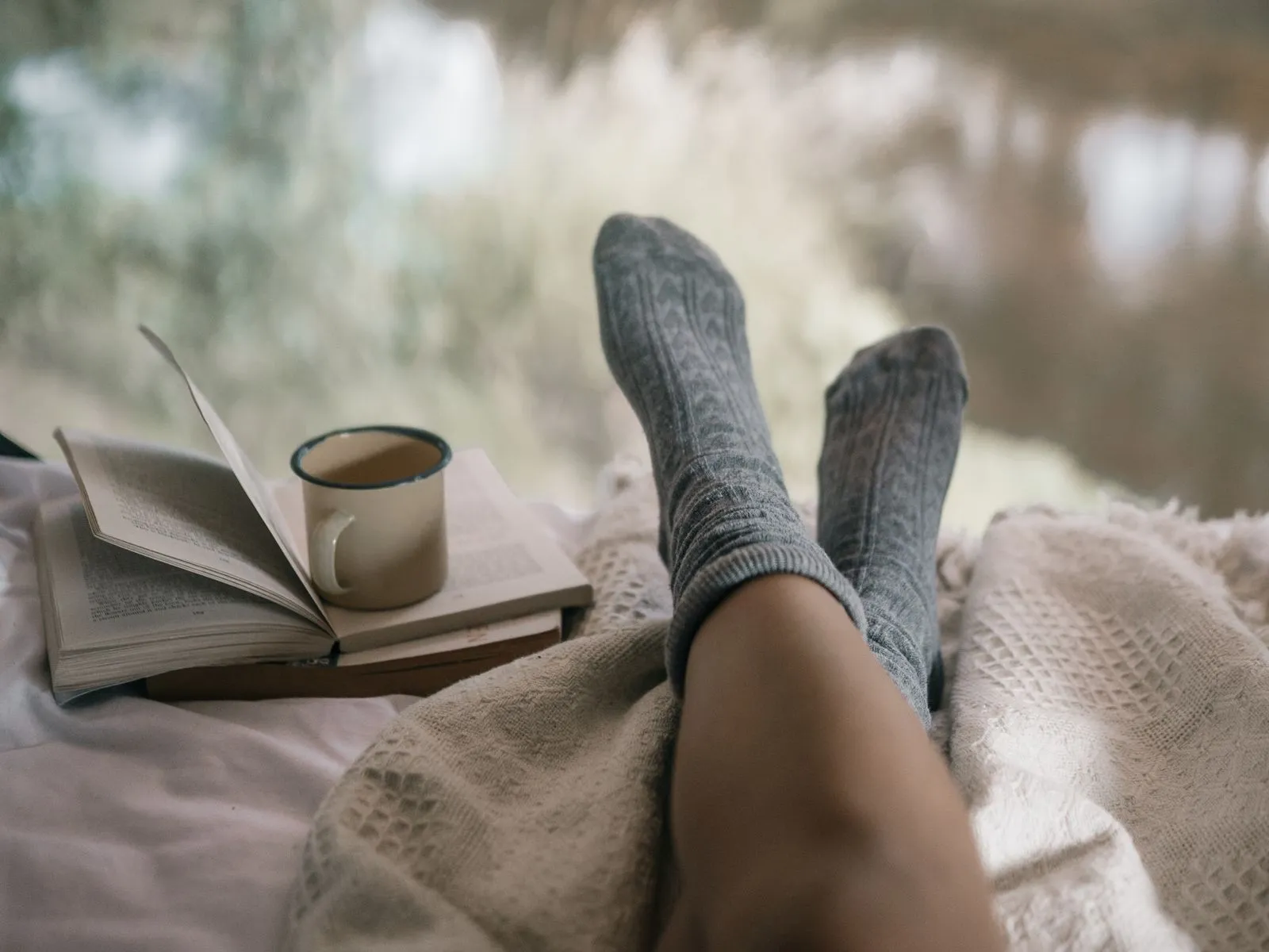 Feet in gray wool socks resting on a cream blanket beside an open book and a mug