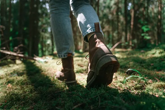 A person's legs and boots mid-stride on a sunlit forest trail